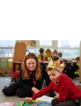 Adult supporting a pupil during a floor-based classroom activity, with the pupil wearing a paper crown and engaging with learning materials.